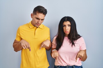Fototapeta premium Young couple standing over isolated background pointing down looking sad and upset, indicating direction with fingers, unhappy and depressed.