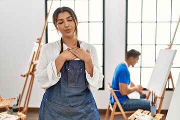 Young hispanic couple at art studio smiling with hands on chest with closed eyes and grateful gesture on face. health concept.