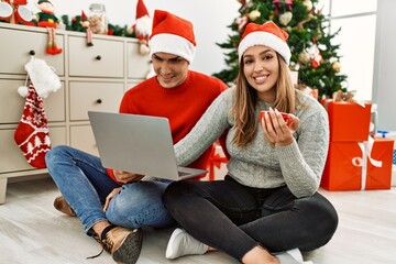 Young couple smiling happy wearing christmas hat. Sitting on the floor using laptop and drinking coffe at home.