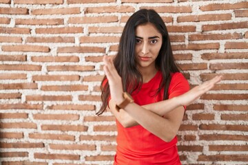 Young teenager girl standing over bricks wall rejection expression crossing arms doing negative sign, angry face