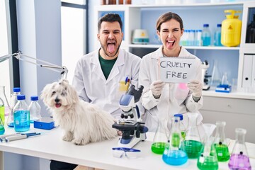 Young hispanic people working at scientist laboratory with dog sticking tongue out happy with funny expression.