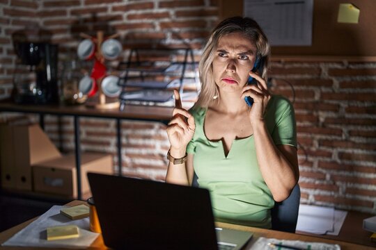 Young Beautiful Woman Working At The Office At Night Speaking On The Phone Pointing Up Looking Sad And Upset, Indicating Direction With Fingers, Unhappy And Depressed.