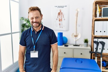 Middle age physiotherapist man working at pain recovery clinic winking looking at the camera with...