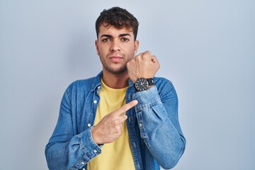 Young hispanic man standing over blue background in hurry pointing to watch time, impatience,...
