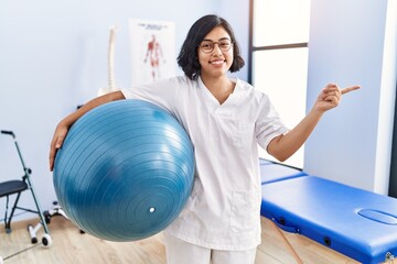 Fototapeta premium Young hispanic physiotherapist woman holding pilates ball smiling happy pointing with hand and finger to the side
