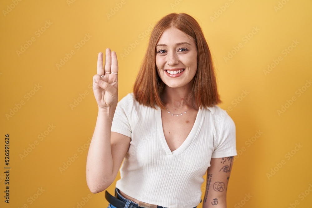 Poster Young redhead woman standing over yellow background showing and pointing up with fingers number three while smiling confident and happy.