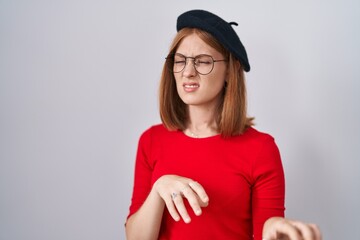 Young redhead woman standing wearing glasses and beret disgusted expression, displeased and fearful doing disgust face because aversion reaction.