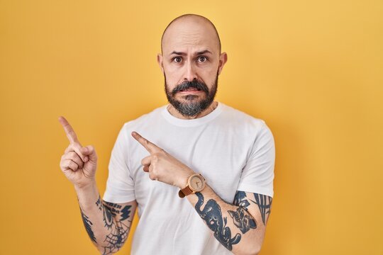Young hispanic man with tattoos standing over yellow background pointing aside worried and nervous with both hands, concerned and surprised expression