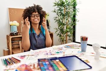 Beautiful african american woman with afro hair painting at art studio gesturing finger crossed smiling with hope and eyes closed. luck and superstitious concept.