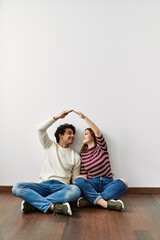 Young hispanic couple doing roof symbol with hands sitting on the floor at empty new home.