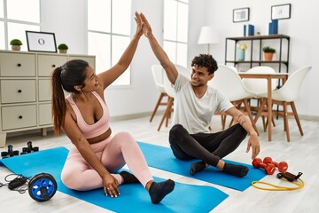 Young latin couple smiling happy training at home. Sitting on the floor high five.