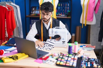 Young hispanic man tailor using sewing machine and laptop at sewing studio
