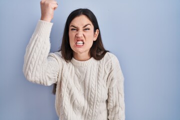Young brunette woman standing over blue background angry and mad raising fist frustrated and...
