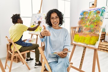 Young hispanic woman at art studio smiling cheerful offering palm hand giving assistance and acceptance.