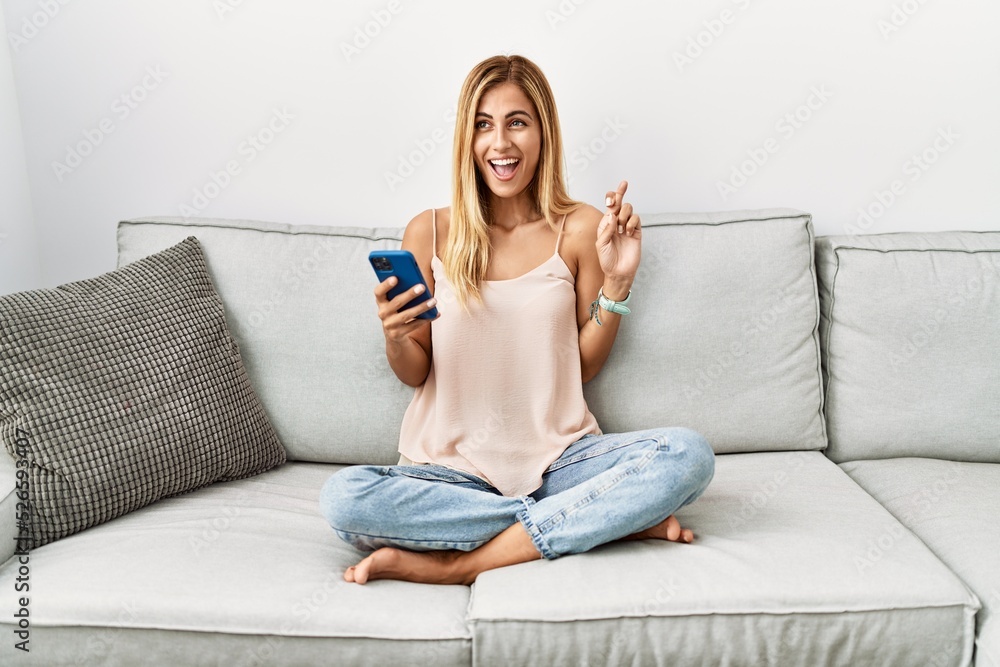 Sticker Blonde beautiful young woman sitting on the sofa at home using smartphone gesturing finger crossed smiling with hope and eyes closed. luck and superstitious concept.