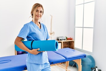 Young caucasian physio therapist smiling happy holding yoga mat at the clinic