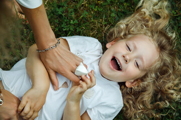 Top view of laughing little girl with long fluffy curly fair hair lying on grass tickled by woman...