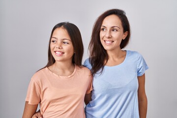 Young mother and daughter standing over white background looking away to side with smile on face, natural expression. laughing confident.