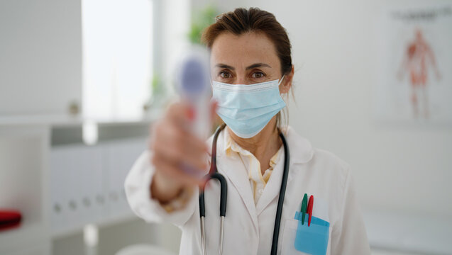 Middle age hispanic woman wearing doctor uniform and medical mask using thermometer at clinic