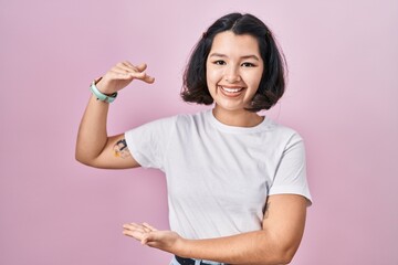 Young hispanic woman wearing casual white t shirt over pink background gesturing with hands showing big and large size sign, measure symbol. smiling looking at the camera. measuring concept.