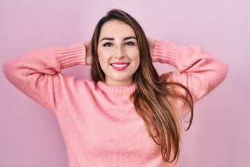 Young hispanic woman standing over pink background relaxing and stretching, arms and hands behind head and neck smiling happy