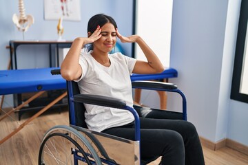 Fototapeta na wymiar Young hispanic woman sitting on wheelchair at physiotherapy clinic with hand on head, headache because stress. suffering migraine.