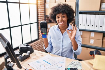 Black woman with curly hair working at small business ecommerce holding credit card and dataphone doing ok sign with fingers, smiling friendly gesturing excellent symbol