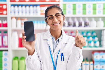 Young hispanic woman working at pharmacy drugstore showing smartphone screen smiling happy and...