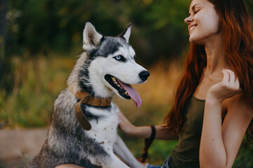 A woman with a husky breed dog smiles and affectionately strokes her beloved dog while walking in nature in the park in autumn against the backdrop of sunset