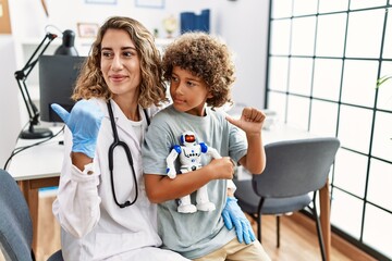 Young kid at pediatrician clinic holding teddy bear pointing thumb up to the side smiling happy with open mouth