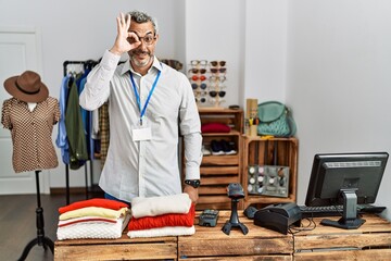 Middle age hispanic man working as manager at retail boutique doing ok gesture with hand smiling, eye looking through fingers with happy face.