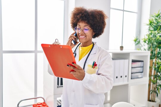 African american woman wearing doctor uniform talking on smartphone reading document at clinic