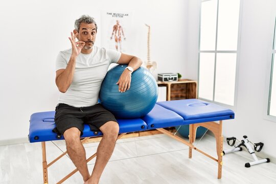 Middle age hispanic man at pain recovery clinic holding pilates ball mouth and lips shut as zip with fingers. secret and silent, taboo talking