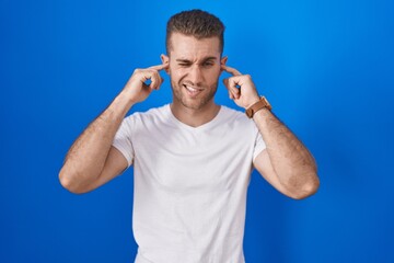 Fototapeta premium Young caucasian man standing over blue background covering ears with fingers with annoyed expression for the noise of loud music. deaf concept.