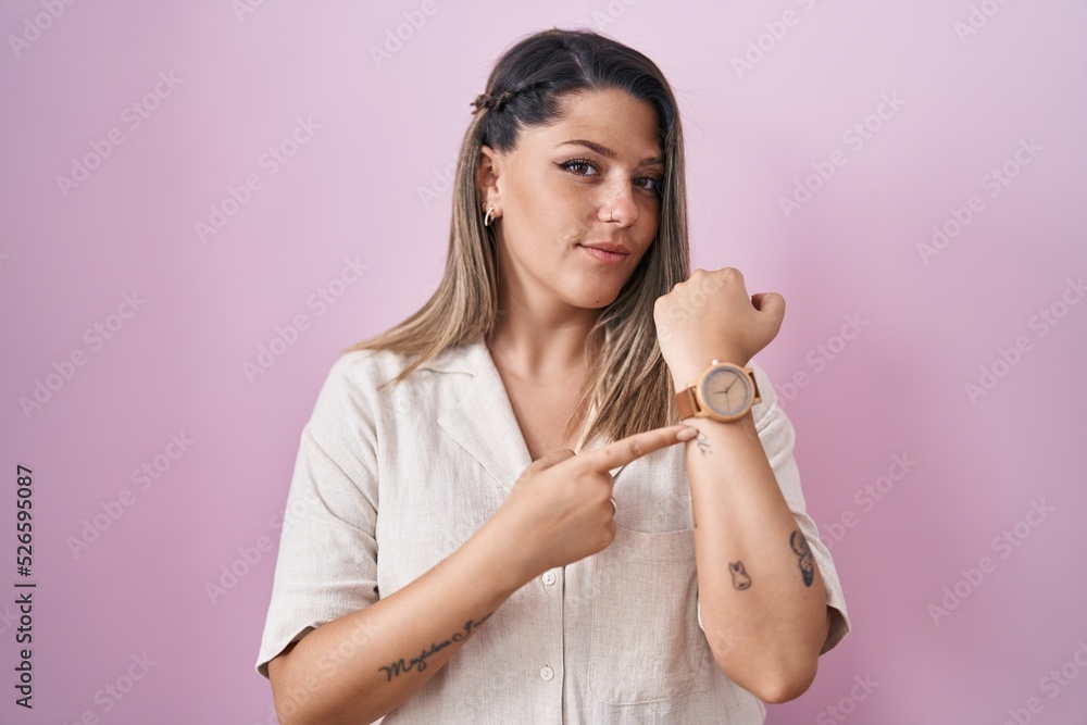 Wall mural Blonde woman standing over pink background in hurry pointing to watch time, impatience, looking at the camera with relaxed expression