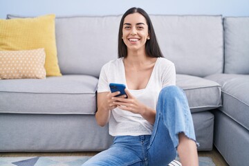 Young hispanic woman using smartphone sitting on floor at home