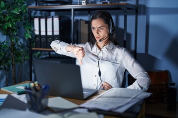 Fototapeta na wymiar Young brunette woman wearing call center agent headset working late at night looking unhappy and angry showing rejection and negative with thumbs down gesture. bad expression.