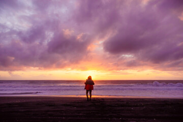 Fototapeta na wymiar silueta de persona masculina parada en la orilla de la playa mirando al atardecer mientras el sol se oculta en el mar con nubes