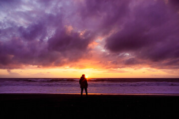 silueta de persona masculina parada en la orilla de la playa mirando al atardecer mientras el sol se oculta en el mar con nubes - obrazy, fototapety, plakaty