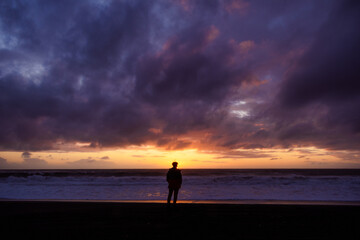silueta de persona masculina parada en la orilla de la playa mirando al atardecer mientras el sol se oculta en el mar con nubes - obrazy, fototapety, plakaty
