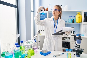 Young blonde woman wearing scientist uniform holding test tube and clipboard at laboratory