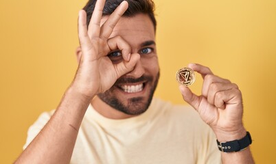 Handsome hispanic man holding tron cryptocurrency coin smiling happy doing ok sign with hand on eye...