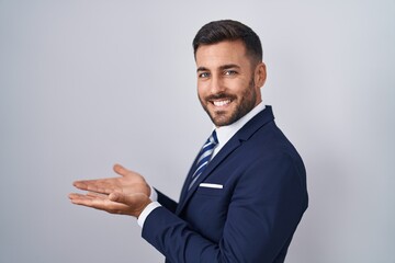 Handsome hispanic man wearing suit and tie pointing aside with hands open palms showing copy space, presenting advertisement smiling excited happy
