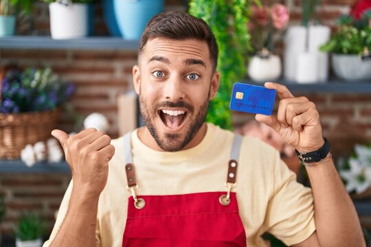 Handsome hispanic man working at florist shop holding credit card pointing thumb up to the side smiling happy with open mouth