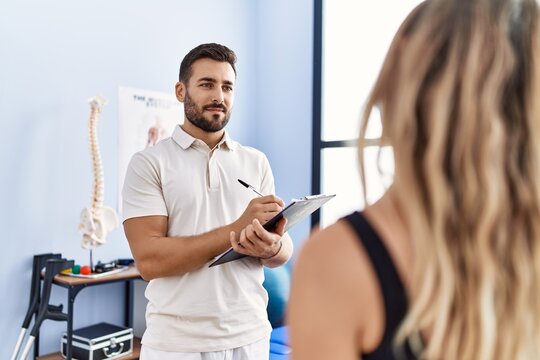 Young hispanic man wearing physiotherapist uniform writing on clipboard at clinic
