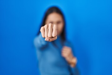 Young brazilian woman standing over blue isolated background punching fist to fight, aggressive and...