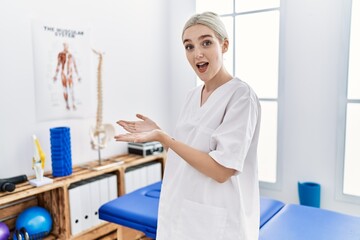 Young caucasian woman working at pain recovery clinic pointing aside with hands open palms showing copy space, presenting advertisement smiling excited happy