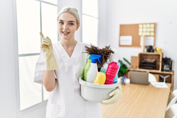 Fototapeta na wymiar Young caucasian woman wearing cleaner uniform holding cleaning products cleaning office smiling happy pointing with hand and finger to the side