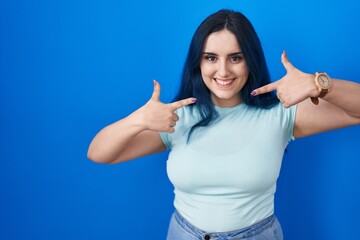 Fototapeta premium Young modern girl with blue hair standing over blue background smiling cheerful showing and pointing with fingers teeth and mouth. dental health concept.