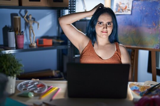 Young modern girl with blue hair sitting at art studio with laptop at night confuse and wonder about question. uncertain with doubt, thinking with hand on head. pensive concept.